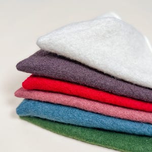 May include: A stack of six rectangular felt fabric pieces in various colors, including white, dark purple, bright red, dusty pink, teal blue, and forest green, arranged in a neat pile with visible textured fibers on each layer.