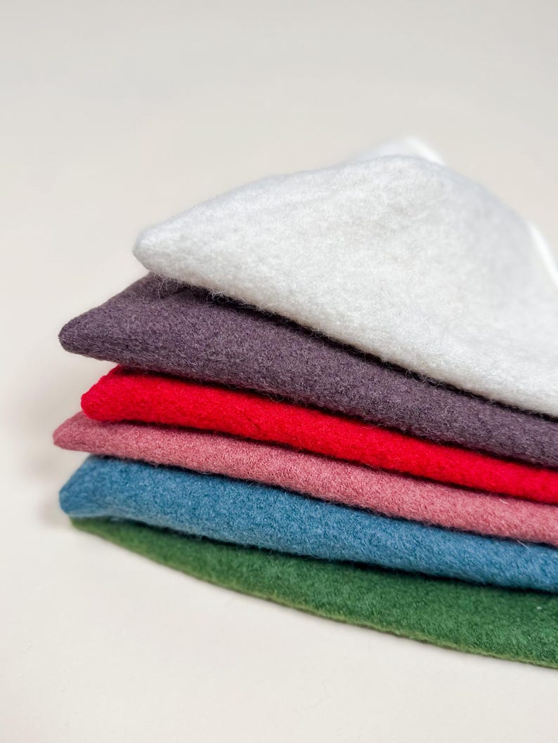May include: A stack of six rectangular felt fabric pieces in various colors, including white, dark purple, bright red, dusty pink, teal blue, and forest green, arranged in a neat pile with visible textured fibers on each layer.