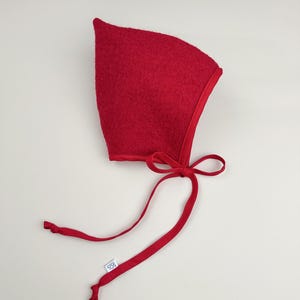 Cranberry boiled wool pixie bonnet with matching cotton ties, handmade and laid flat on a neutral background.