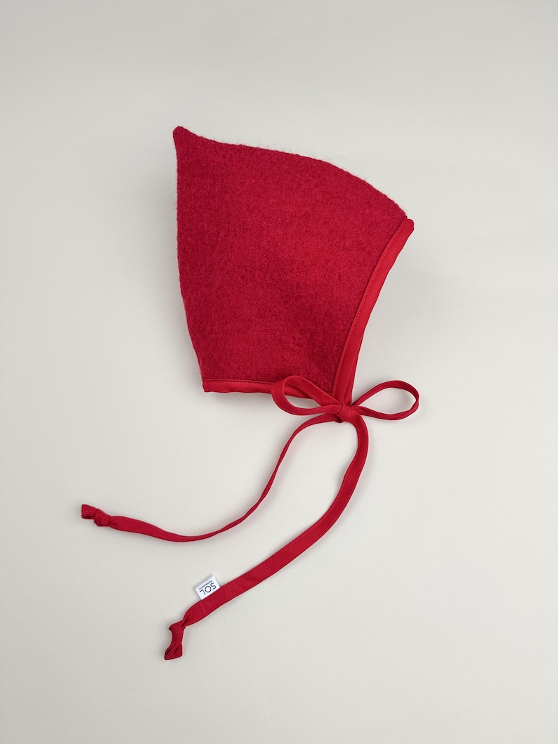 Cranberry boiled wool pixie bonnet with matching cotton ties, handmade and laid flat on a neutral background.