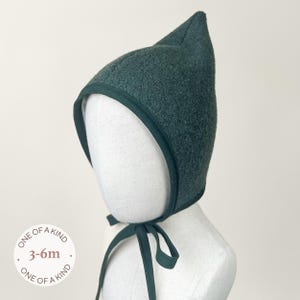 May include: A dark green baby bonnet with a pointed top and a contrasting trim. The bonnet has a tie closure and is designed for babies aged 3-6 months. The text "ONE OF A KIND" is printed on the image.