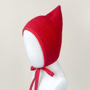 Red boiled wool pixie bonnet with pointed crown and matching cotton ties, displayed on a linen mannequin against a soft neutral background.