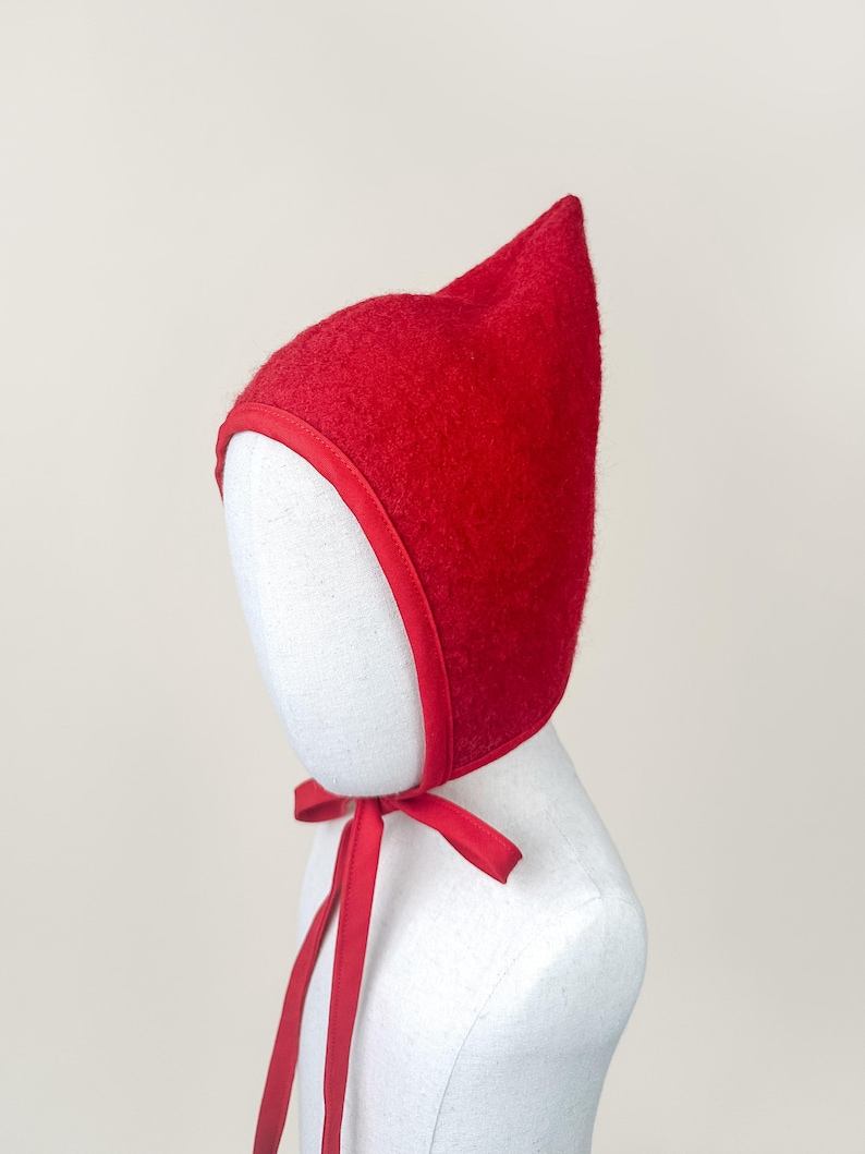 Red boiled wool pixie bonnet with pointed crown and matching cotton ties, displayed on a linen mannequin against a soft neutral background.