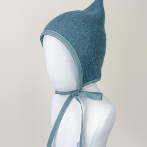 Juniper boiled wool pixie bonnet modeled on a white mannequin, showing front angle and tied bow.
