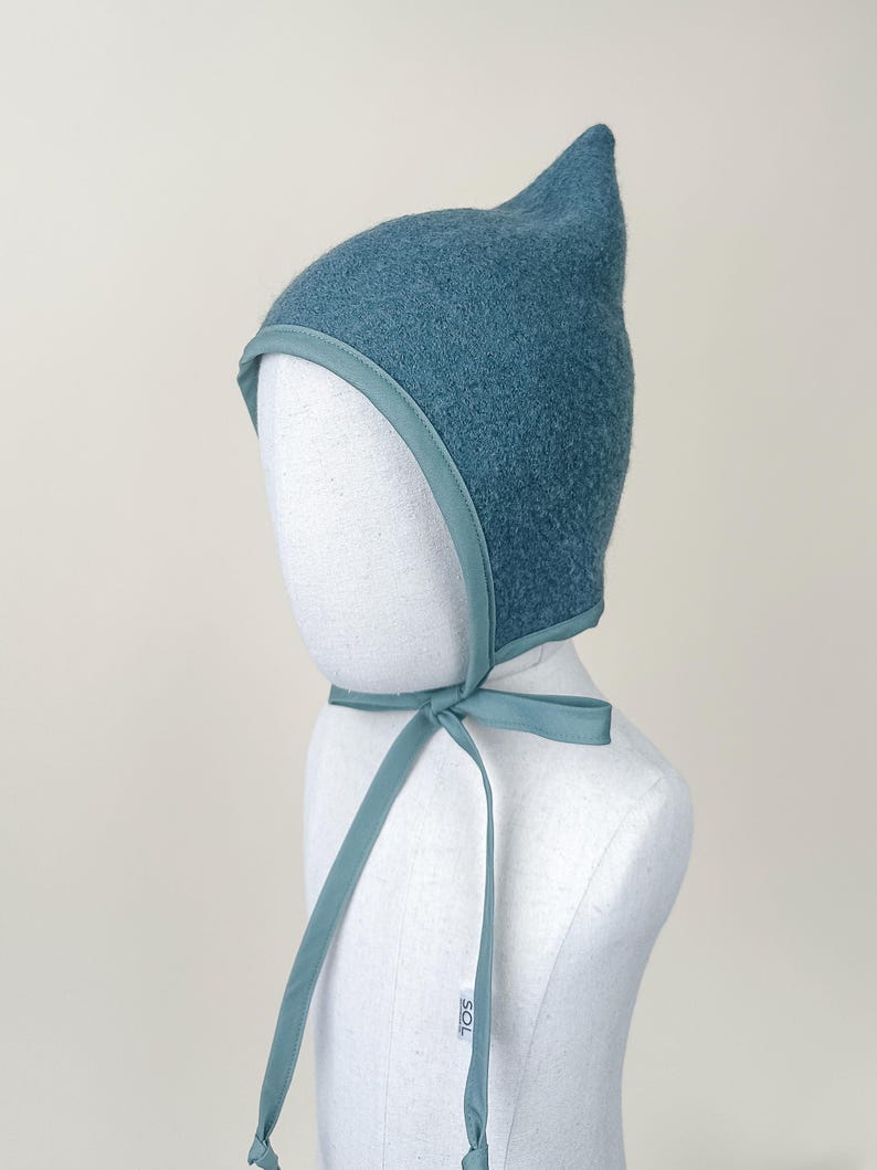 Juniper boiled wool pixie bonnet modeled on a white mannequin, showing front angle and tied bow.