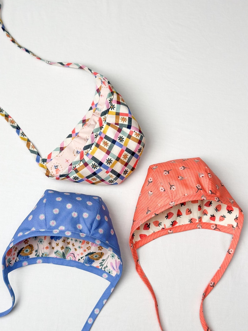 Three reversible baby & toddler bonnets with different prints, including coral floral and strawberry designs, laid flat.