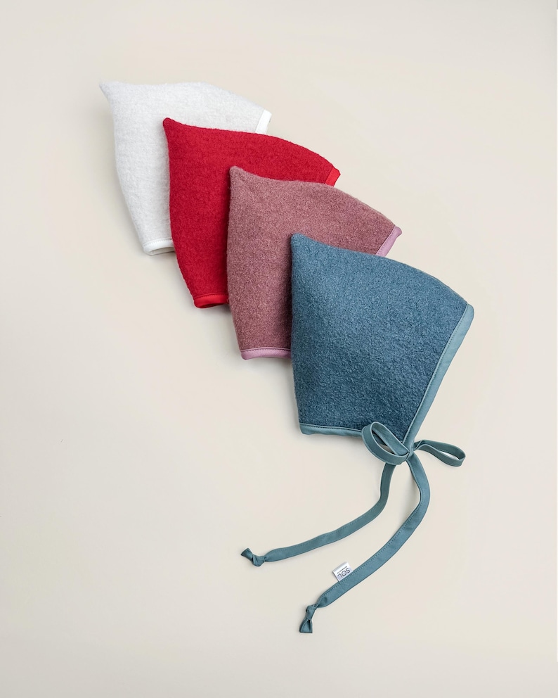Four boiled wool pixie bonnets in cozy winter shades — Snowdrop (ivory), Cranberry (red), Rosewood (rose), and Juniper (green) — displayed flat on a cream background.