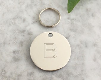 May include: Silver round dog tag with a letter B engraved on it. The tag is attached to a silver key ring.