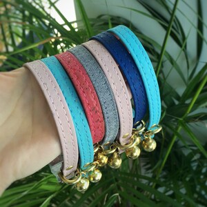 May include: A set of five colorful cat collars with bells. The collars are made of different materials, including pink, blue, and red fabric, cork, and denim. The collars are all decorated with gold-colored bells.