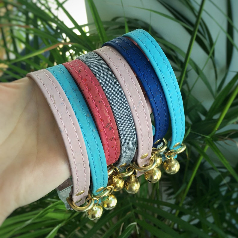 May include: A set of five colorful cat collars with bells. The collars are made of different materials, including pink, blue, and red fabric, cork, and denim. The collars are all decorated with gold-colored bells.