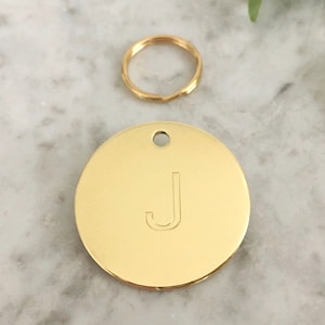 May include: Gold-coloured metal dog tag with the letter J engraved on the front. The tag is attached to a gold-coloured metal key ring.