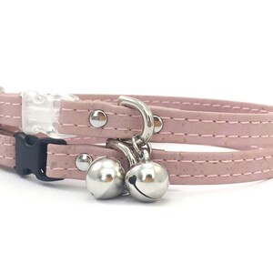 May include: A pink cat collar with a breakaway buckle and two silver bells. The collar is made of cork and has a stitched design.