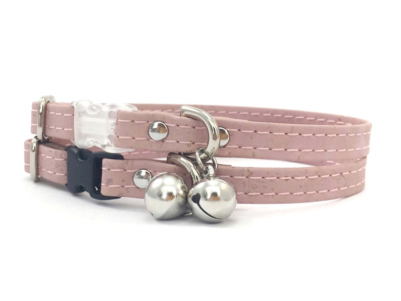May include: A pink cat collar with a breakaway buckle and two silver bells. The collar is made of cork and has a stitched design.