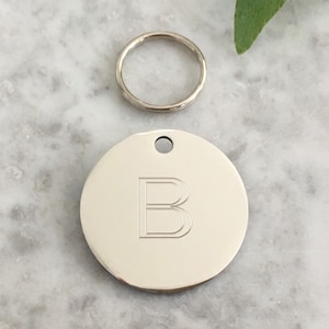 May include: Silver round dog tag with a letter B engraved on it. The tag is attached to a silver key ring.