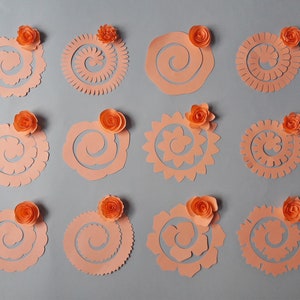 May include: An assortment of peach-colored paper flower cutouts in various spiral and floral designs. Each cutout is topped with a small, orange paper rose. The cutouts are arranged on a gray background, showcasing the different shapes and textures.