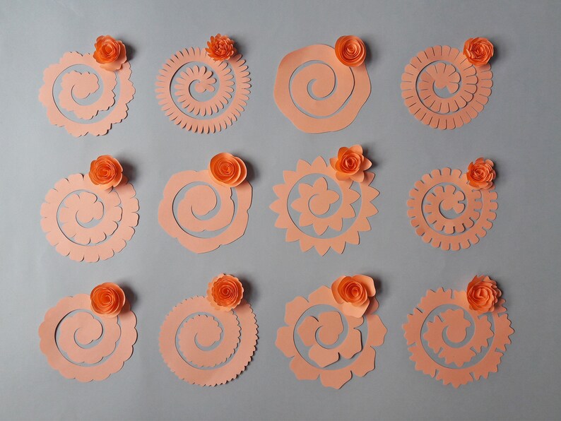 May include: An assortment of peach-colored paper flower cutouts in various spiral and floral designs. Each cutout is topped with a small, orange paper rose. The cutouts are arranged on a gray background, showcasing the different shapes and textures.