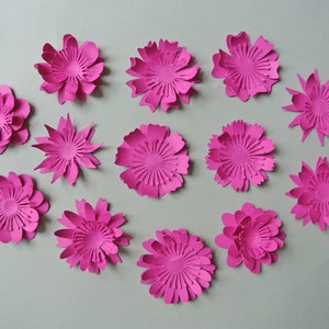 May include: A collection of 13 pink paper flowers, each with a different design and layered petals. The flowers are arranged in a semi-circle on a gray background.