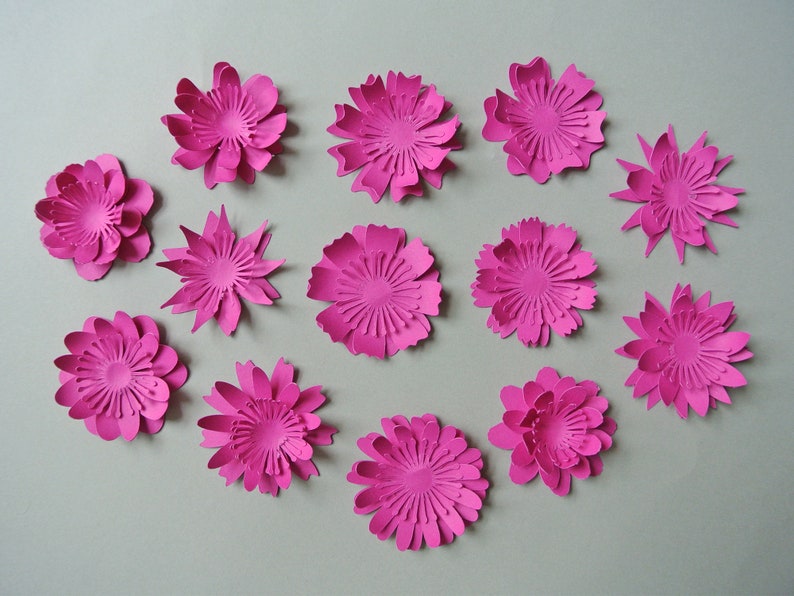 May include: A collection of 13 pink paper flowers, each with a different design and layered petals. The flowers are arranged in a semi-circle on a gray background.