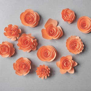May include: An assortment of peach-colored paper flowers in various sizes and styles, arranged on a gray background. The flowers have a layered, rolled design, resembling roses. The arrangement includes both open and tightly wound blooms, creating a visually appealing display.