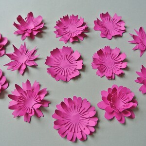 May include: A collection of 13 pink paper flowers, each with a different design and layered petals. The flowers are arranged on a white surface.