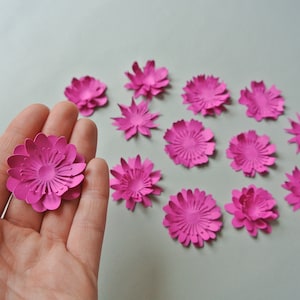 May include: A collection of 13 pink paper flowers, each with a layered, 3D design. The flowers are arranged in a semi-circle on a white surface.
