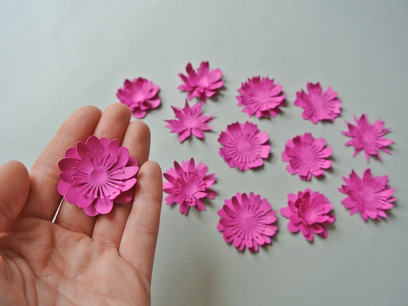 May include: A collection of 13 pink paper flowers, each with a layered, 3D design. The flowers are arranged in a semi-circle on a white surface.