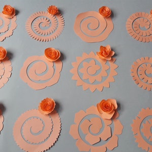 May include: A collection of peach-colored paper cutouts in various spiral and floral designs, each topped with a small, orange paper flower. The cutouts are arranged on a gray background, showcasing the different shapes and textures.