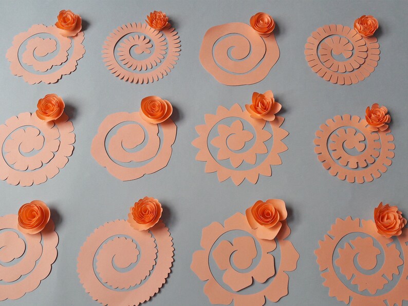 May include: A collection of peach-colored paper cutouts in various spiral and floral designs, each topped with a small, orange paper flower. The cutouts are arranged on a gray background, showcasing the different shapes and textures.
