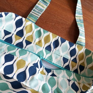 Peut inclure: Un grand sac fourre-tout avec une ouverture zipp&eacute;e en haut. Le sac est fait d'un tissu blanc avec un motif g&eacute;om&eacute;trique bleu, vert et or. Les bretelles sont faites d'un tissu ray&eacute;.