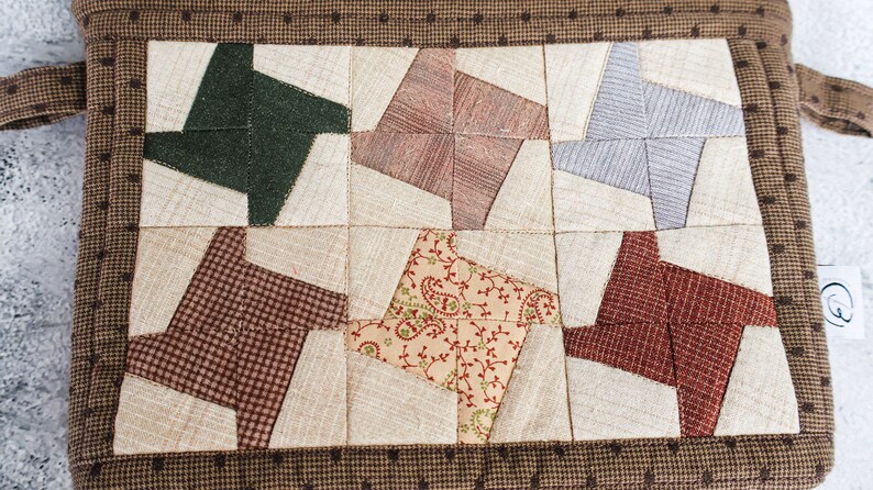 May include: A brown quilted hot pad with a patchwork design featuring six different fabric patterns in shades of brown, green, red, beige, and grey. The hot pad has a brown border with white polka dots.