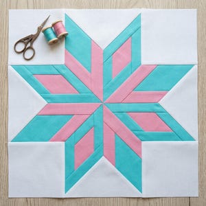 May include: A square quilt block featuring a central eight-pointed star pattern in alternating teal and light pink fabric segments on a white background, accompanied by a pair of metal embroidery scissors and two small spools of thread.