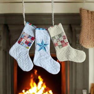 Puede incluir: Tres calcetines navideños colgantes de tela blanca acolchada. Uno tiene un diseño de patchwork, otro una estrella azul y el tercero un diseño de patchwork. Los calcetines están suspendidos frente a una chimenea.