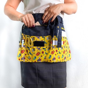 May include: A yellow gardening apron with a black and white striped waistband. The apron has multiple pockets and is decorated with a pattern of colorful bees and flowers.