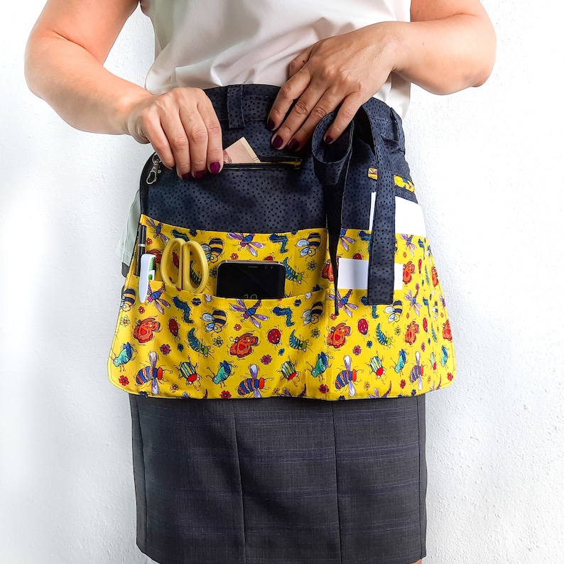 May include: A yellow gardening apron with a black and white striped waistband. The apron has multiple pockets and is decorated with a pattern of colorful bees and flowers.