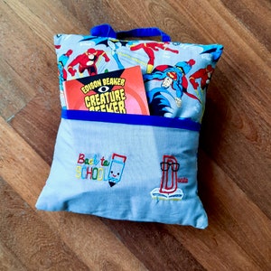 May include: Blue and gray pillow with a pocket on the front. The pocket has a book inside with the title "Edison Beaker Creature Seeker". The pillow has an embroidered design that says "Back to School" with a pencil and a book.