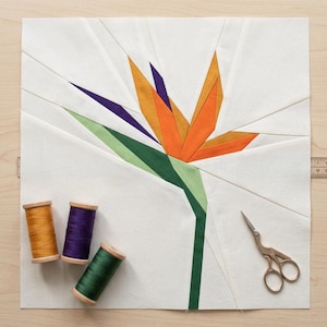 Può includere: Un blocco di tessuto patchwork con un motivo a fiore di uccello del paradiso nei colori arancione, viola e verde. Tre rocchetti di filo in oro, viola e verde e un paio di forbici dorate sono posizionati vicino al blocco. Il disegno è su sfondo bianco.
