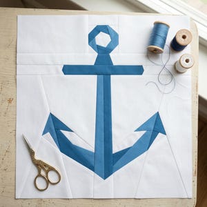 May include: A square fabric quilt block featuring a pieced blue anchor design on a white background. The anchor is constructed from various shades of blue fabric. Gold-colored metal scissors and three wooden spools of blue thread are present.