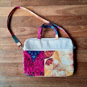 K&ouml;nnte beinhalten: Eine wei&szlig;e Canvas-Laptoptasche mit einem bunten, gemusterten Stofffutter. Das Futter hat ein pinkfarbenes, blaues, gelbes und wei&szlig;es Blumenmuster. Die Tasche hat einen langen, verstellbaren Riemen.