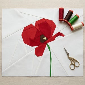 Könnte beinhalten: Ein Stoffkunstwerk mit einer roten Mohnblume mit braunem Zentrum und grünem Stiel auf weißem Hintergrund. Das Kunstwerk wird von Garnrollen in verschiedenen Farben und einer kleinen goldenen Schere begleitet. Die Mohnblume besteht aus geometrischen Formen.