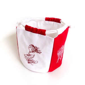 Puede incluir: Una bolsa de cordón blanco y rojo con un diseño bordado blanco de una niña con una flor en la cabeza. La bolsa tiene un cordón blanco y está hecha de una tela suave.
