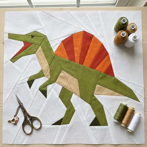May include: A fabric quilt block featuring a pieced Spinosaurus dinosaur in shades of olive green, tan, and orange, set against a white background. Includes metal embroidery scissors with a bird charm and six spools of thread.