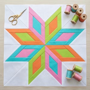 Pode incluir: Um bloco de colcha quadrado com um padrão central de estrela de oito pontas em segmentos de tecido rosa brilhante, turquesa, verde-limão e laranja sobre um fundo branco, acompanhado por tesouras de metal dourado e carretéis de linha de madeira.