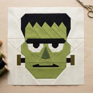 May include: A square fabric quilt block featuring a pieced design of a green-skinned character with black hair, thick black eyebrows, a triangular nose, and a straight black mouth, set against a cream-colored background with geometric seams.