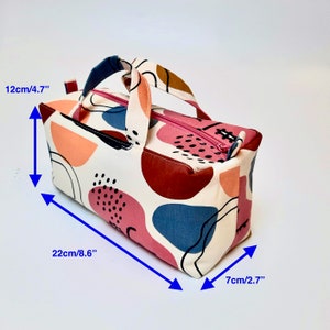 May include: A white fabric tote bag with a pink zipper and a brown handle. The bag has a colorful abstract pattern with pink, red, blue, and orange shapes. The bag measures 8.6 inches long, 4.7 inches tall, and 2.7 inches wide.