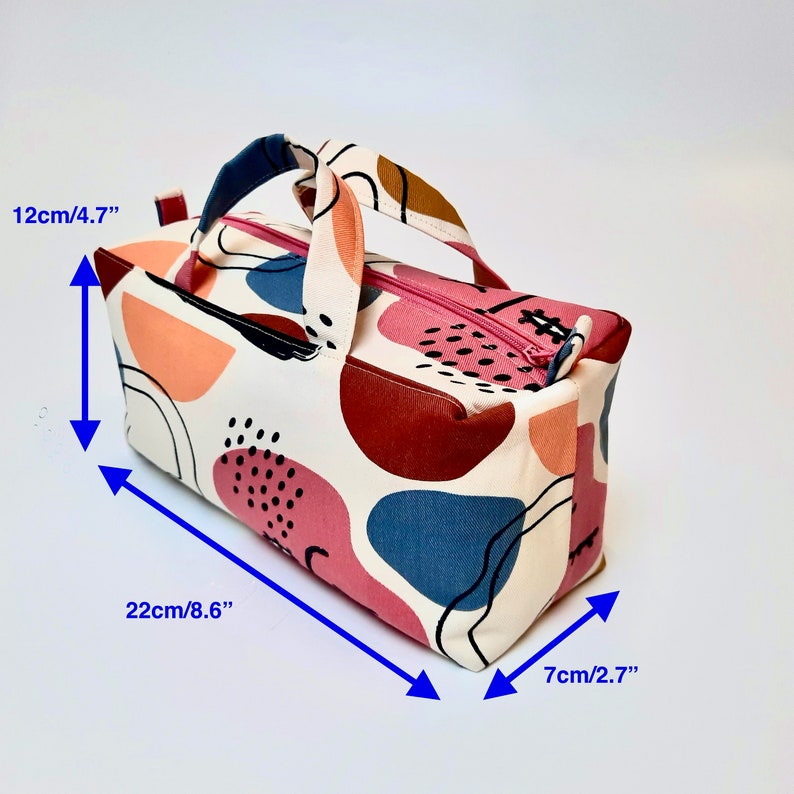 May include: A white fabric tote bag with a pink zipper and a brown handle. The bag has a colorful abstract pattern with pink, red, blue, and orange shapes. The bag measures 8.6 inches long, 4.7 inches tall, and 2.7 inches wide.