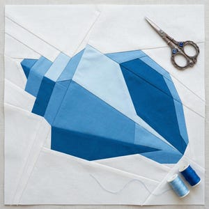 Könnte beinhalten: Ein quadratischer Stoff-Quiltblock mit einem geometrischen Muscheldesign, das aus verschiedenen Blau- und Weißtönen von Baumwollstoff zusammengesetzt ist. Eine verzierte Metallschere und zwei kleine blaue Nähgarnrollen liegen auf dem Block.