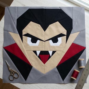 May include: A square fabric quilt block featuring a pieced vampire face design in black, beige, white, red, and grey. The vampire has sharp white fangs, a red tongue, and a high collar. A pair of scissors, two spools of thread, and a ruler are visible.