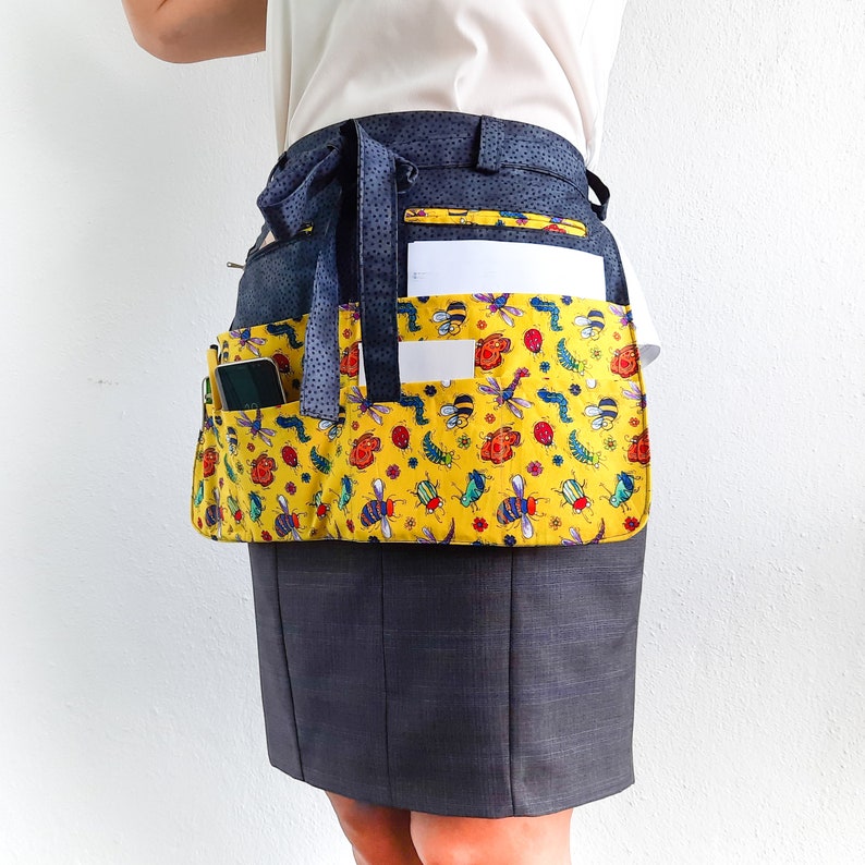 May include: A black and yellow gardening apron with a bee and flower pattern. The apron has multiple pockets, including a large pocket on the front and a smaller pocket on the side. The apron is worn over a white shirt and a pair of dark grey pants.
