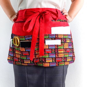 May include: Red apron with a double pocket design and a red ribbon tie. The apron has a colorful bus pattern with pockets for holding tools or supplies.