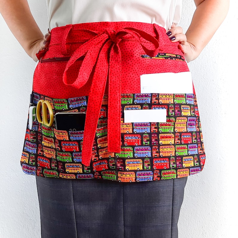 May include: Red apron with a double pocket design and a red ribbon tie. The apron has a colorful bus pattern with pockets for holding tools or supplies.